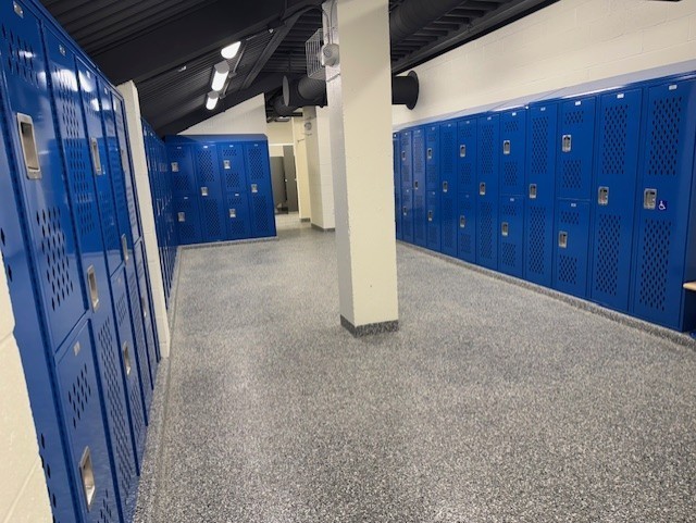 Long hallway with blue lockers on both sides and a central white column, along with a concrete floor and lights on the ceiling.