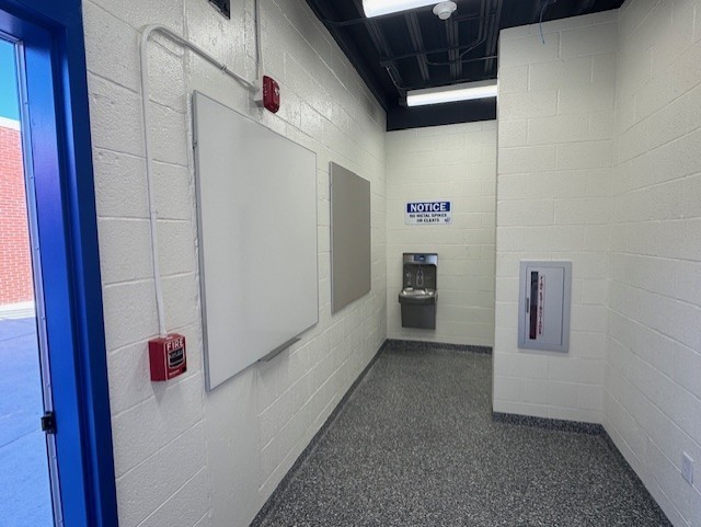 An empty room with white walls and gray flooring. A whiteboard is mounted on the wall. A red box is attached to the wall.