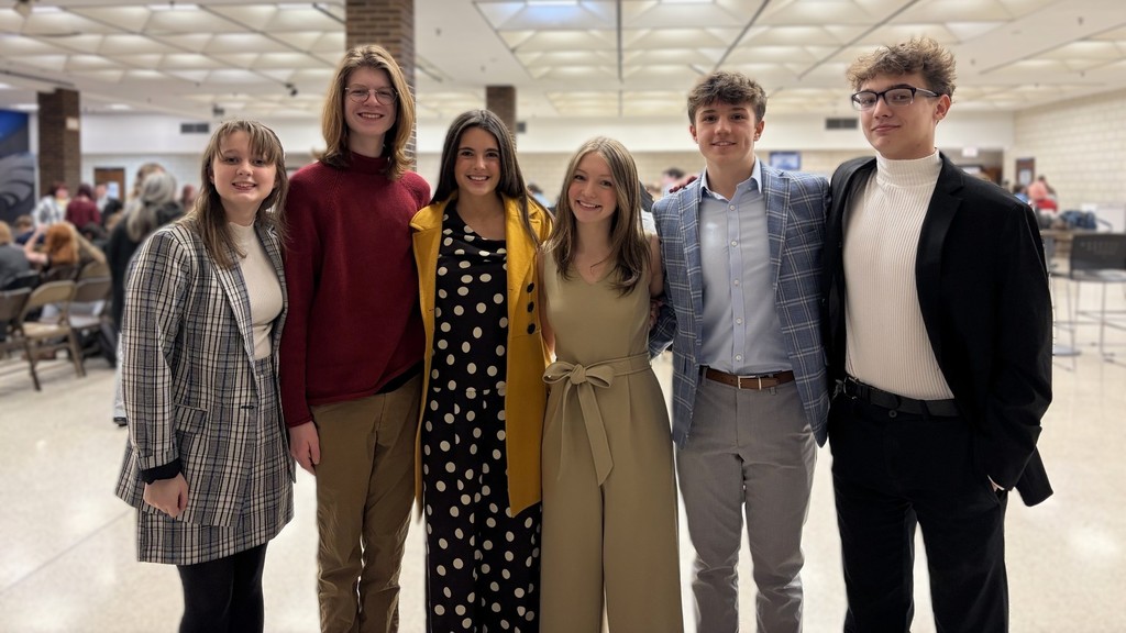 Six people in winter clothing pose together in a room with a white ceiling and large windows.