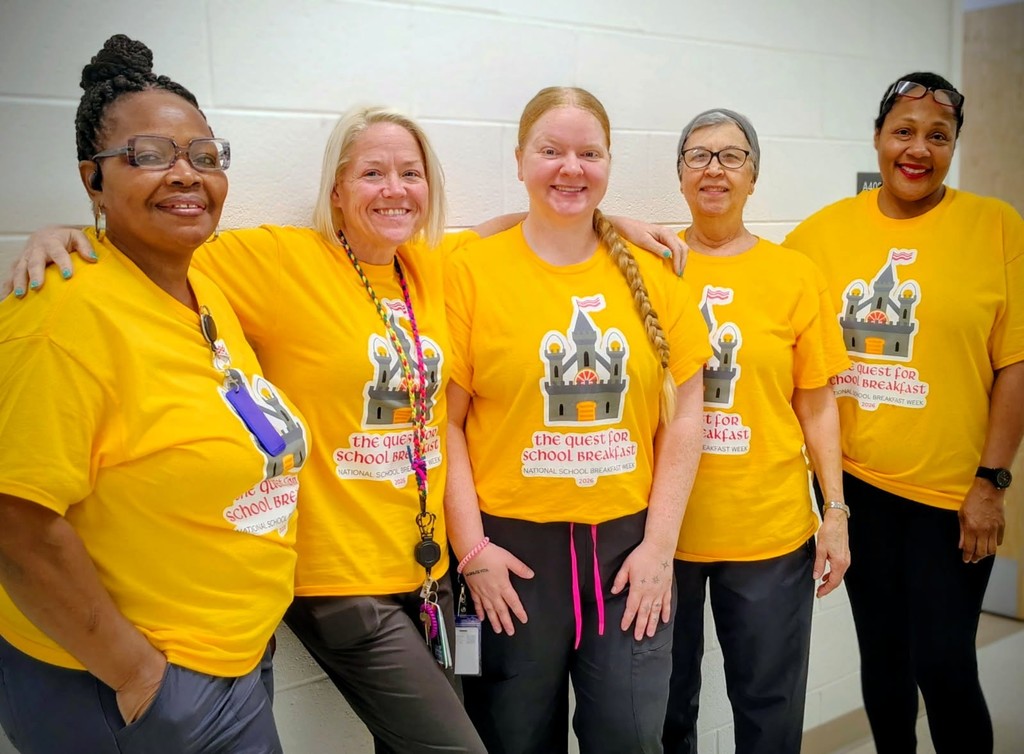 Five women wearing yellow shirts with cartoon designs stand close together, smiling, and posing for a photo.