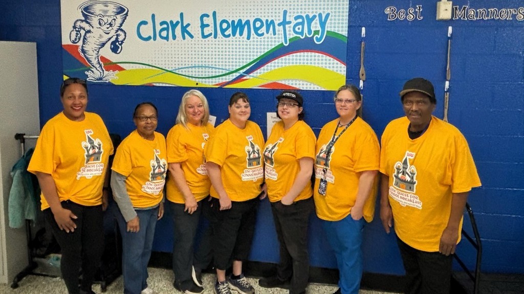 A group of adults wearing yellow shirts and hats pose in front of a blue wall with a banner.