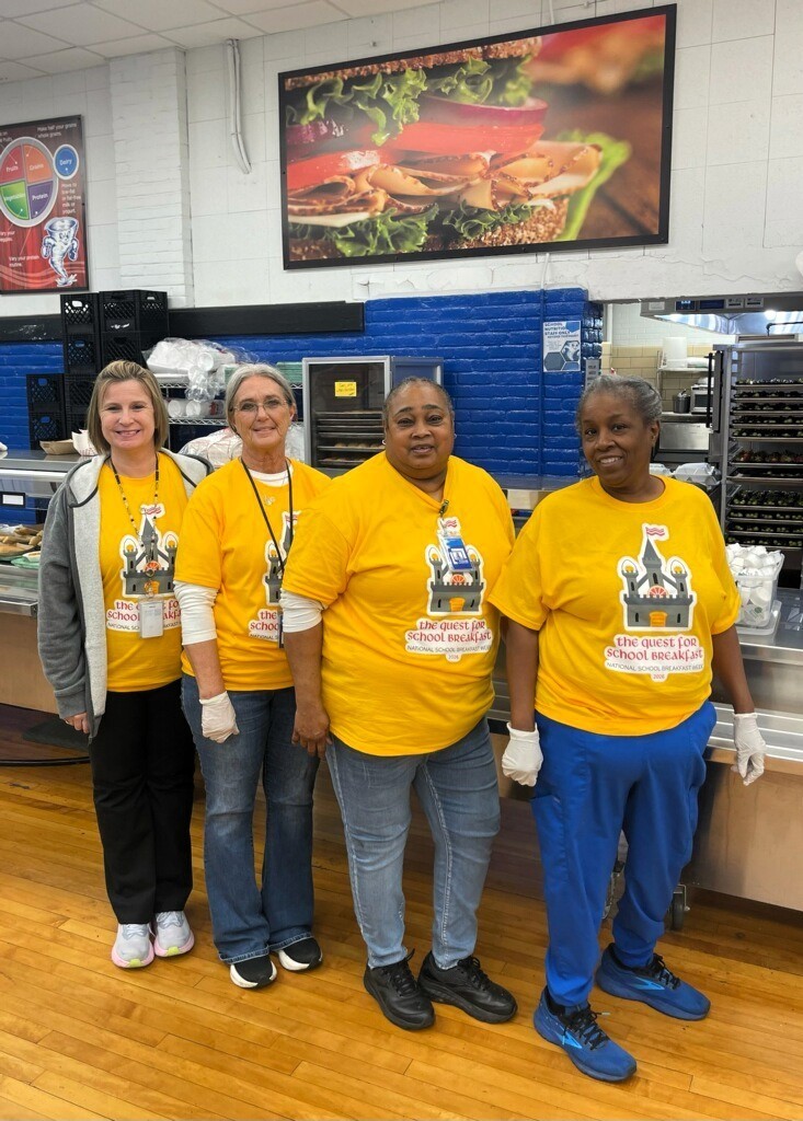 Four individuals wearing yellow shirts, white gloves, and sneakers stand together in a kitchen. A counter and shelves are behind them.