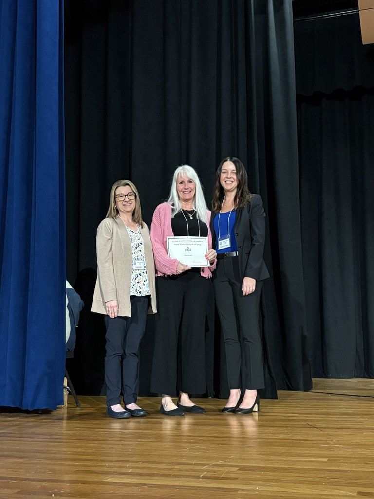 Three women stand on a stage, one holding a certificate. The setting is a stage with a blue curtain in the background.