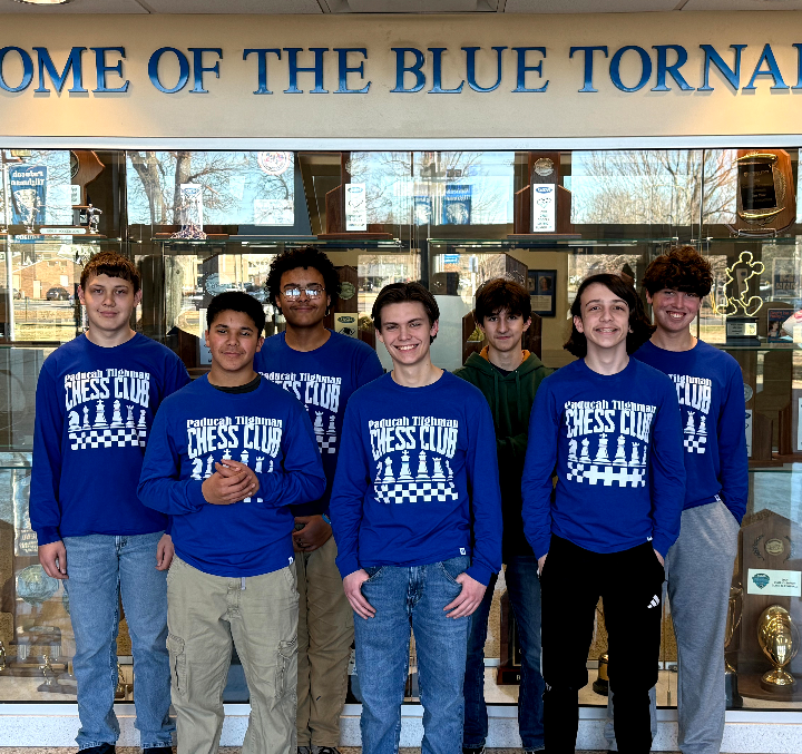 A group of young men in matching blue shirts stand in front of a chess club sign.
