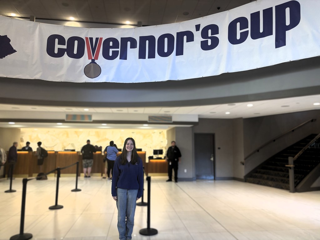A woman stands in a lobby with a banner reading "Governor's Cup" above her. She is in front of a check-in desk.