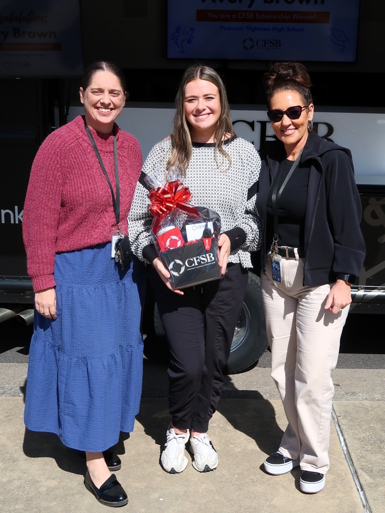 Three women stand together on a sidewalk, with one holding a gift box. The background includes a bus.