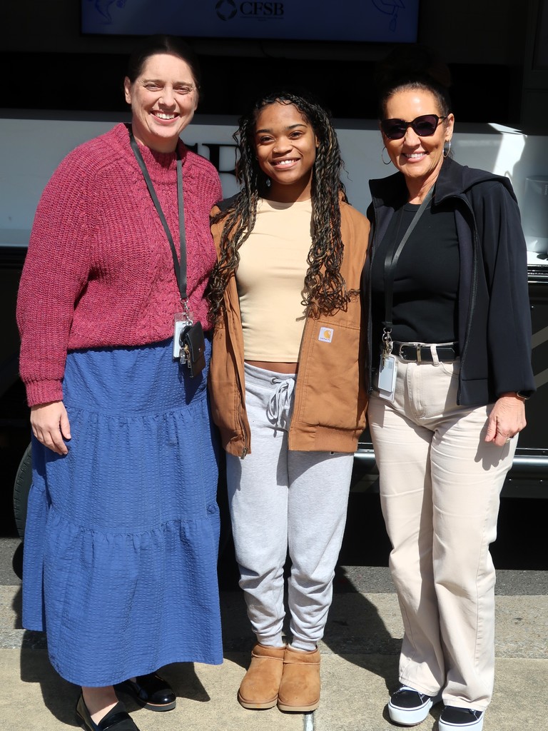 Three women pose for a photo outdoors, with one wearing a pink sweater, another in a brown jacket, and the last in a black jacket.