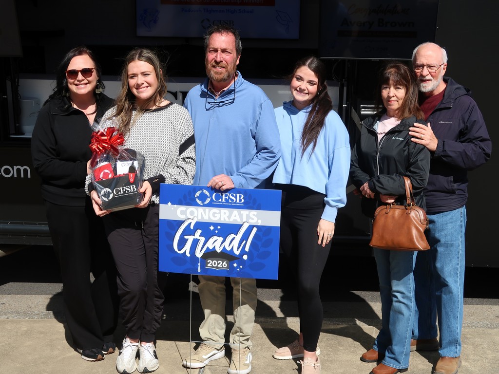 A group of six people in casual clothing stand together outdoors. A man holds a blue sign with "CONGRATS Grad!" text.