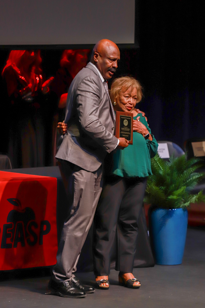 A person on stage with an award, holding it up while hugging another person. Behind them, a red banner with letters.
