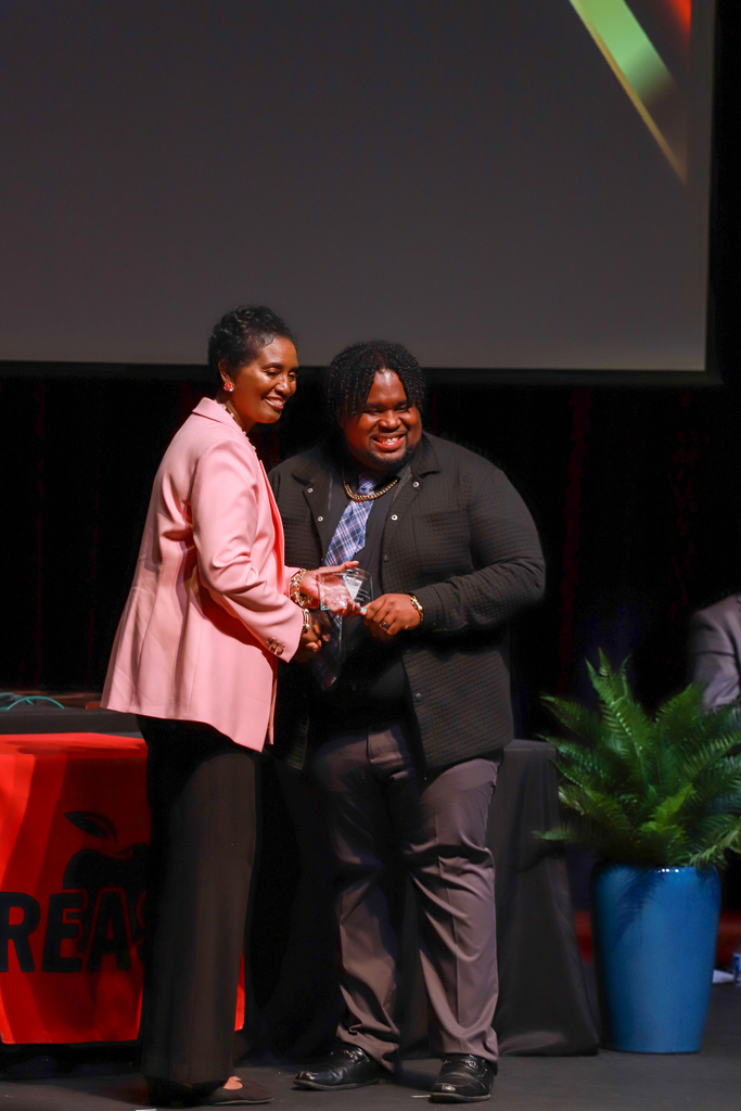 A person in a black suit accepts an award from a person in a pink jacket on a stage.