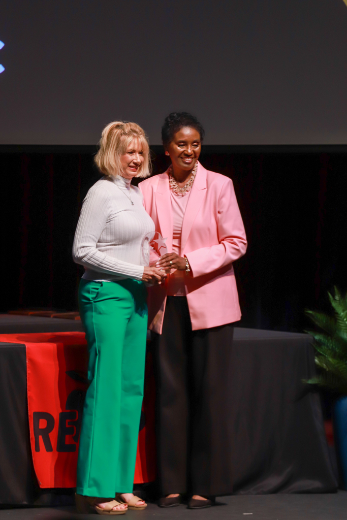 Two women stand on stage with one holding a trophy. They are smiling and shaking hands. Behind them, a red table displays a sign.