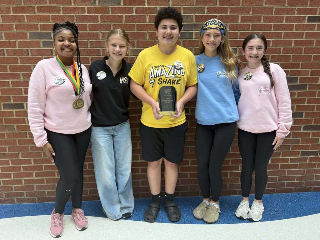 Five individuals, one holding a trophy, stand close together in front of a brick wall. They wear various colored shirts and pants.