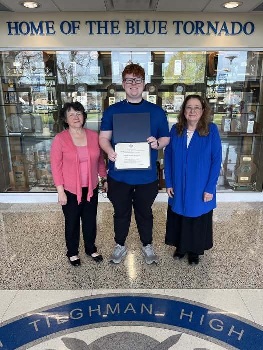 Three individuals pose in front of a building. The man in the center holds a certificate. Two women stand beside him.