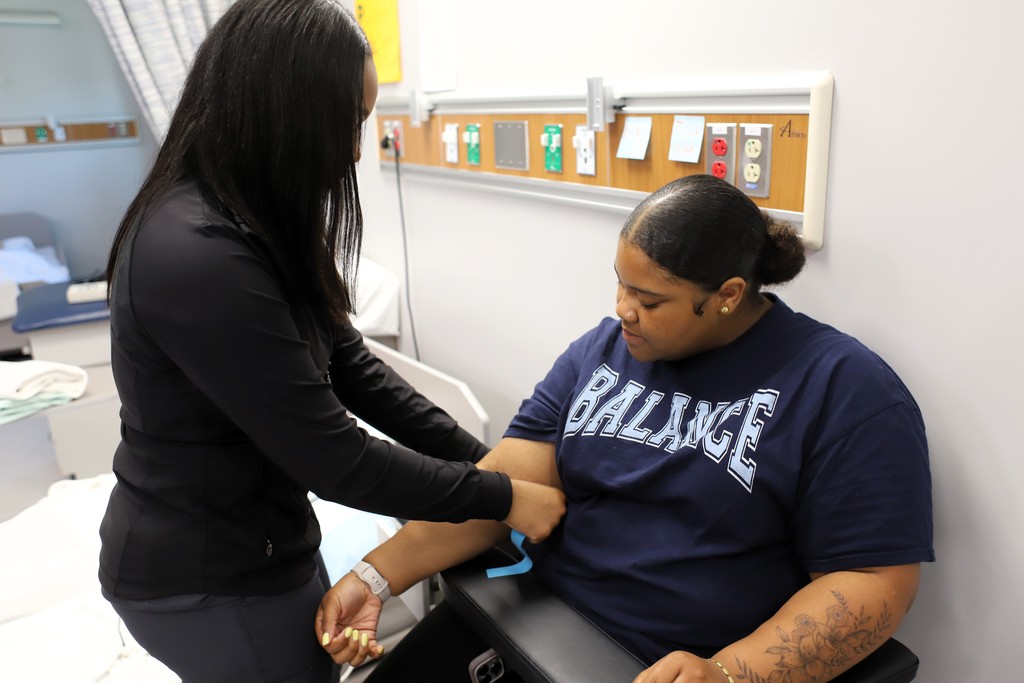 A person is seated in a hospital chair while another person, in blue, takes their blood.