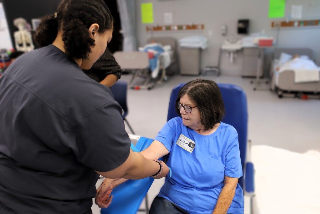 Two women in a hospital room. One is seated in a chair, another stands beside her, adjusting a cuff.