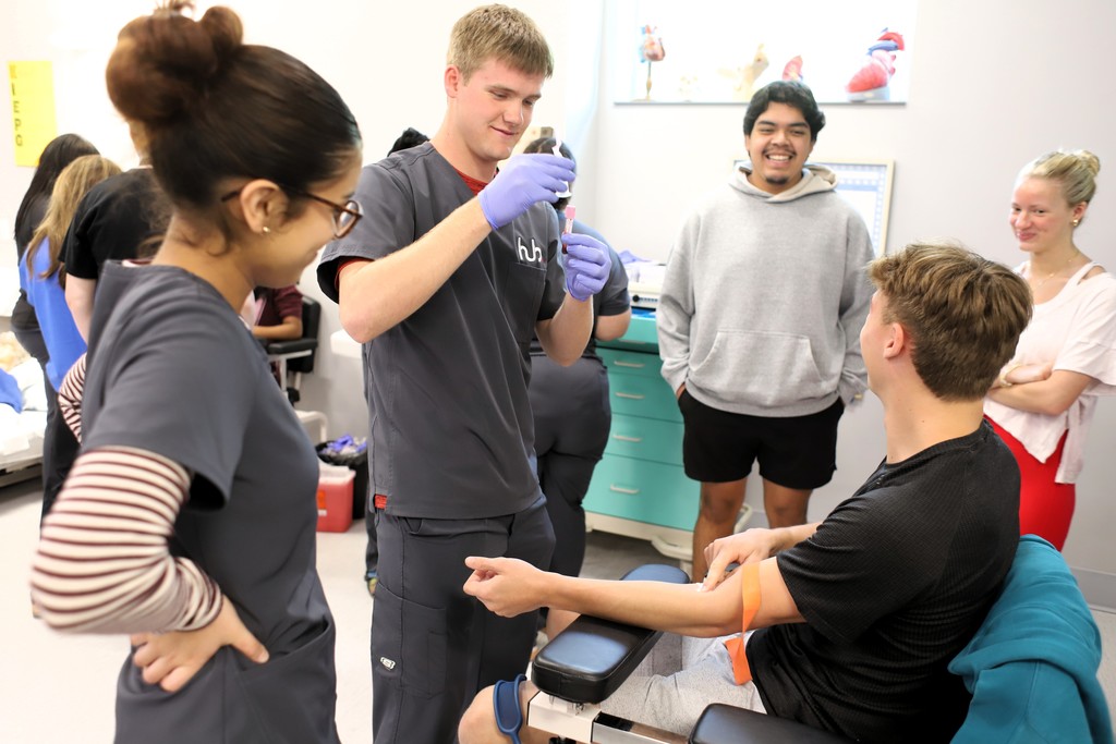 A group of people in medical attire, one in gloves preparing an injection, another seated receiving it, and others observing in a clinical room.