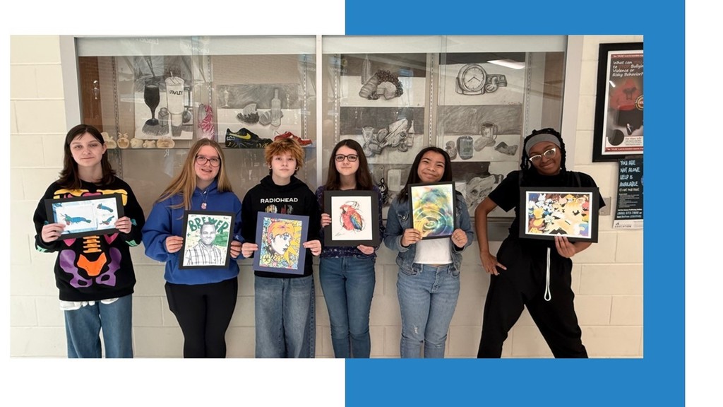 Seven individuals, dressed in various outfits, stand in a row and hold framed artworks. A white wall with multiple framed artworks is in the background.