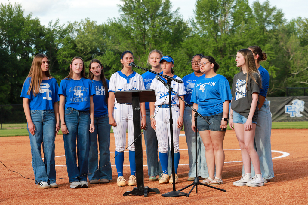 Nine people stand on a baseball field, wearing blue shirts and jeans. One holds a microphone.