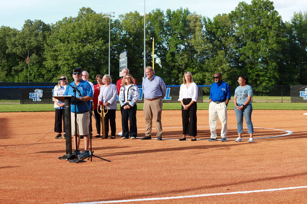 A group of people stand on a baseball field with one person speaking at a podium.