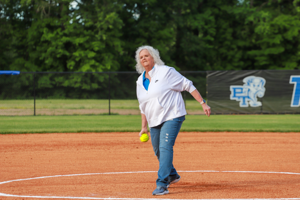 An adult woman with white hair pitches a softball on a field with a white circle.