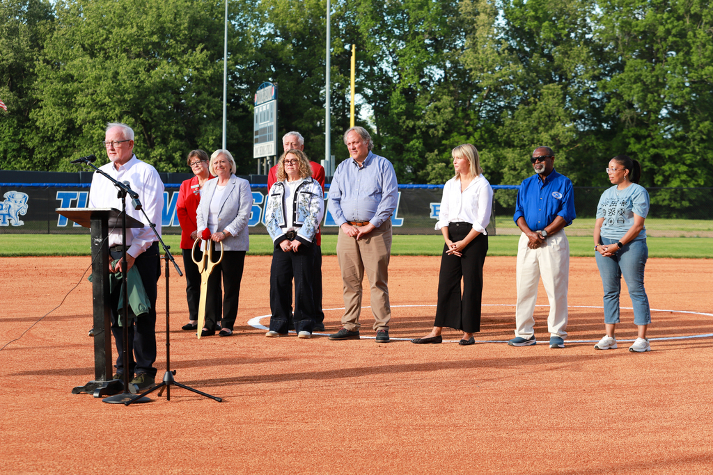 A group of adults stand on a baseball field for a ribbon cutting ceremony. A microphone is set up at a podium.
