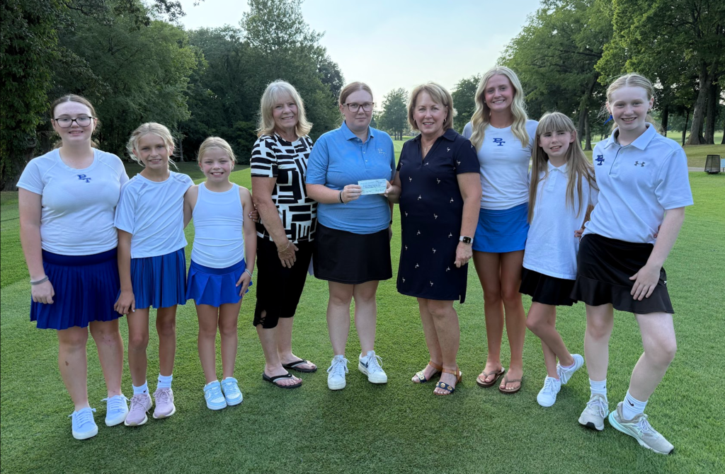 Nine women and girls in matching shirts and skirts pose on a golf course, with a large check in the middle.