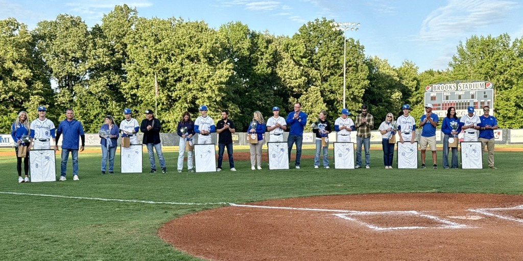 Group of people in blue and white shirts standing on a baseball field with trees in the background.