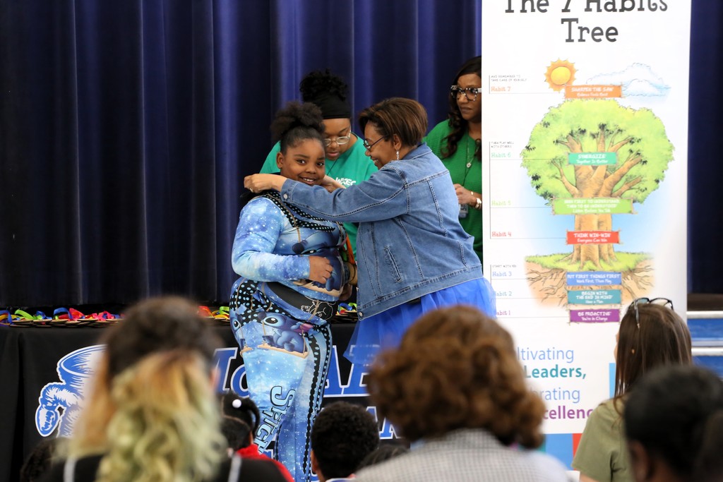 An adult and a child are standing before a crowd. The adult is giving the child a hug.
