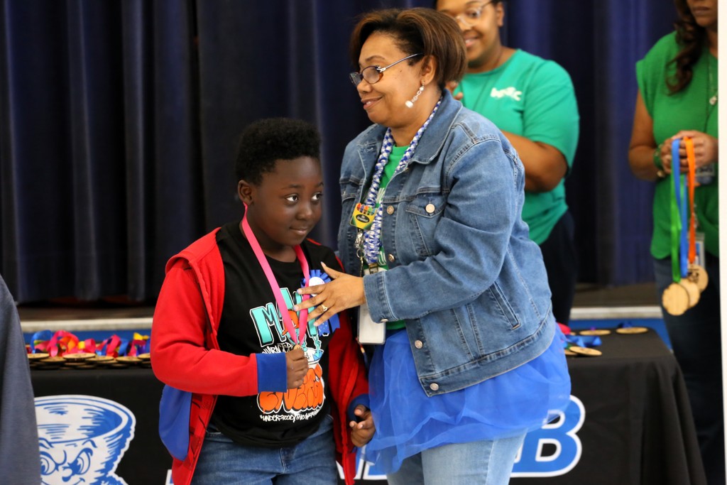 Two people in a room, one holding a medal, standing before a table with a banner.