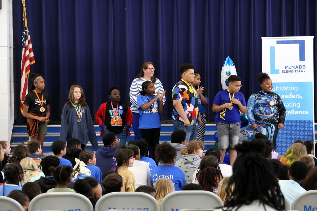 Group of young people in blue shirts standing on a stage, some wearing medals. Audience seated in front.