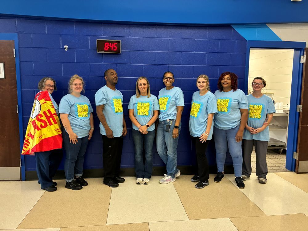 Group of people in matching light blue shirts stand in a hallway; some hold a flag.