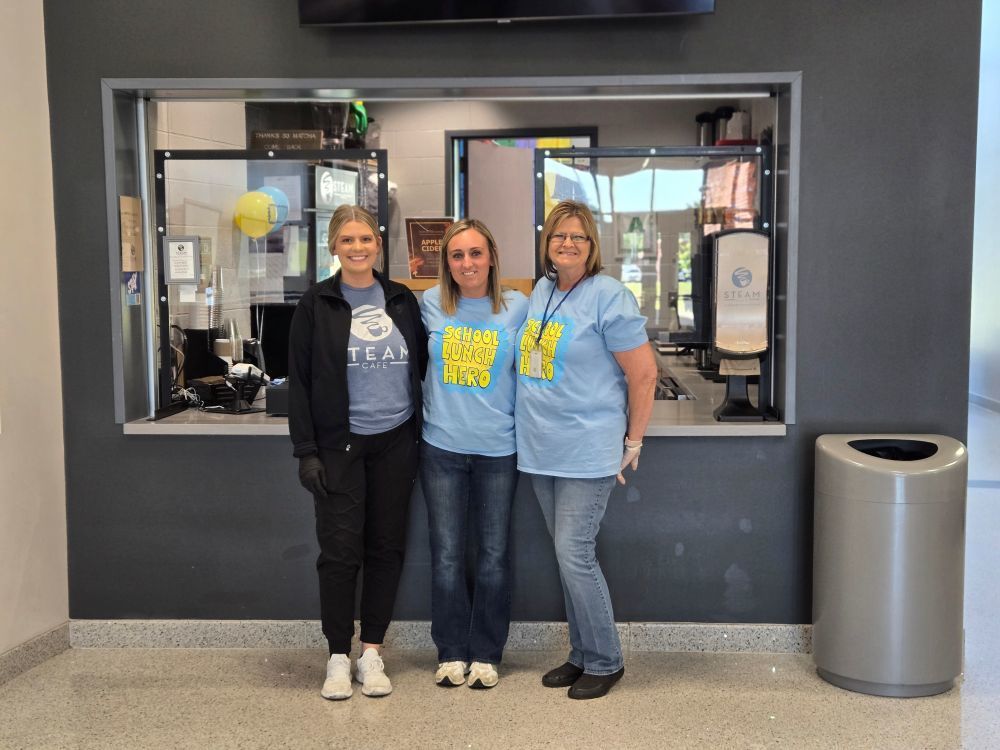 Three women stand together in front of a glass window with an office interior. Two wear blue shirts, one wears a black jacket.