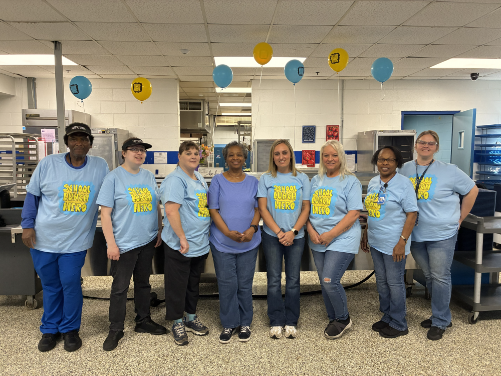 A group of seven people in light blue shirts stand in a room with white walls, balloons, and kitchen equipment.