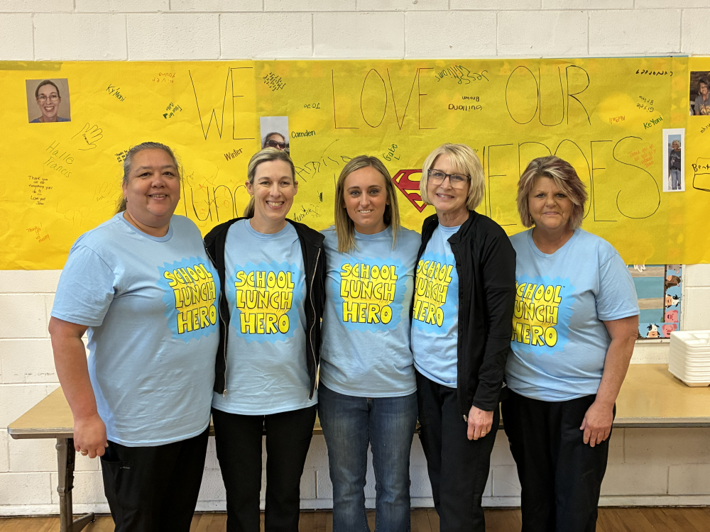 Five women in matching light blue shirts stand together. They face the camera. A yellow banner with "We Love Our Heroes" is behind them.