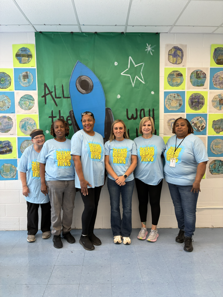 Six individuals in matching blue shirts and sneakers pose in front of a green banner with a rocket and star.