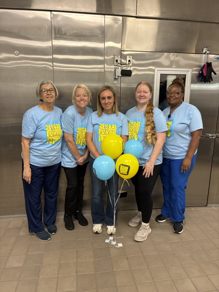 Five women in matching light blue shirts stand close together with balloons. One holds a bouquet. Tile floor and a white door behind them.