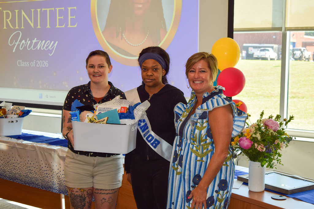 Three women stand with a box of gifts, one wearing a sash, and a presentation behind them.