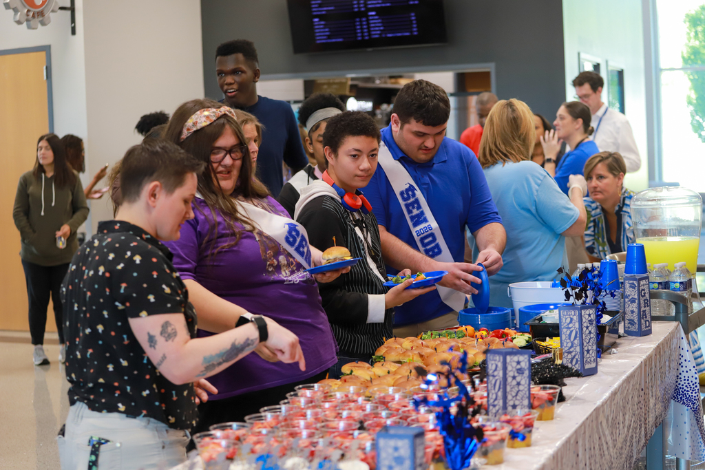 People gather around a table with food, drinks, and decorations. Some are wearing sashes, and others are interacting.