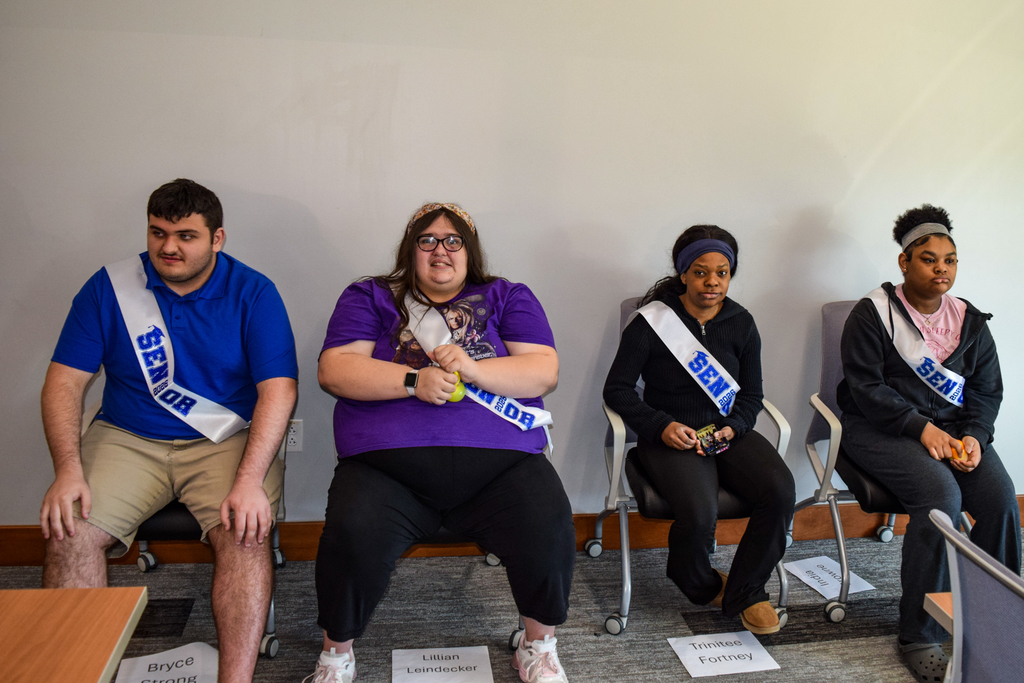 Four individuals sit in chairs wearing sashes, two white, one black, and one pink. One wears a blue shirt.