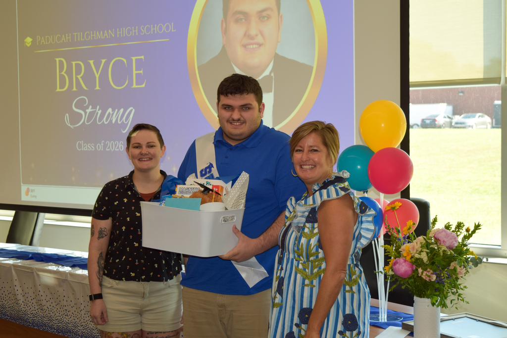 Three individuals, two women and a man, stand in front of a projector screen. A bouquet and balloons are in the foreground.