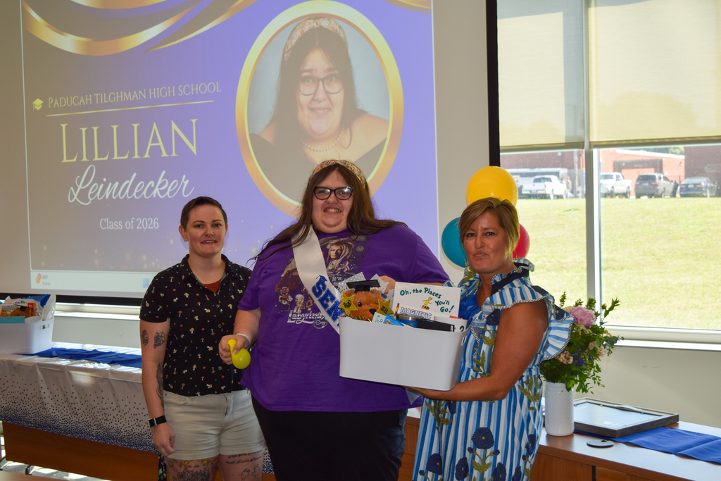 Three women pose for a photo; one holds a box of gifts, another a ball, and one stands in front of a projector screen.