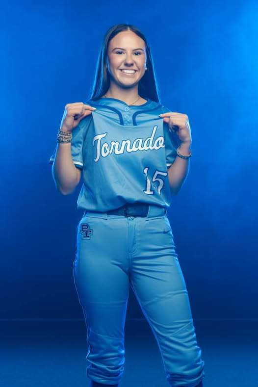 A woman in a teal uniform with "Tornado" and number 15 holds up her jersey.
