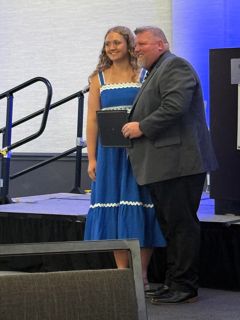 A man and a woman stand on a stage, smiling. The woman holds an award. A black railing is behind them.