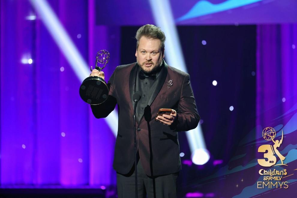 A man in a black suit holds an award under stage lights, with purple background and spotlights.