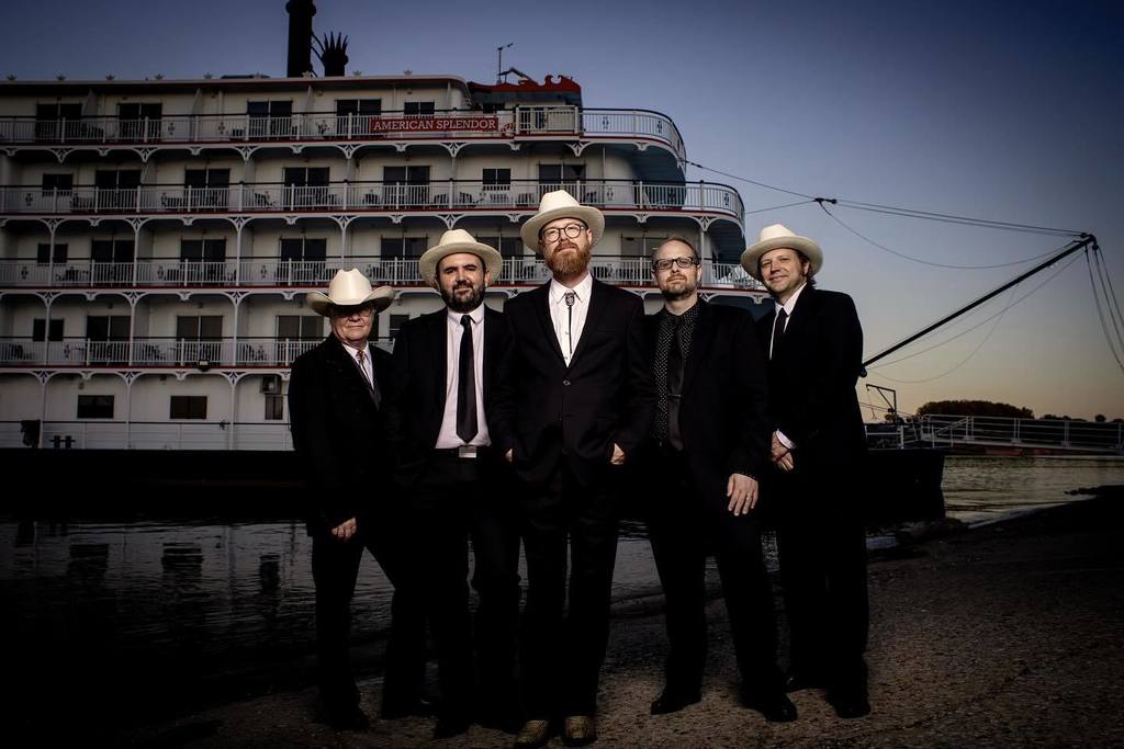 Five men in suits and hats stand together in front of a riverboat.