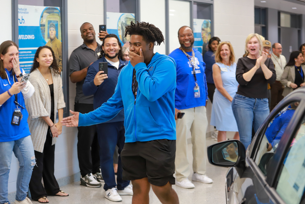 A group of people, some holding phones, watch a man in a blue jacket and black shorts walk by.