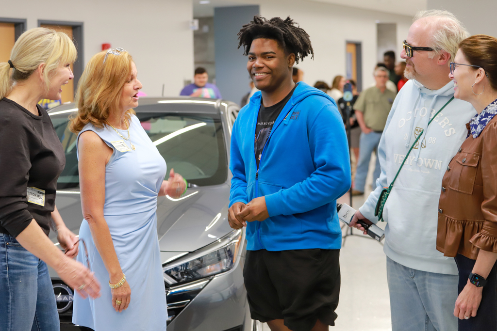 A group of people stands around a car in a hall. A man in a blue hoodie smiles, while others wear casual clothing.