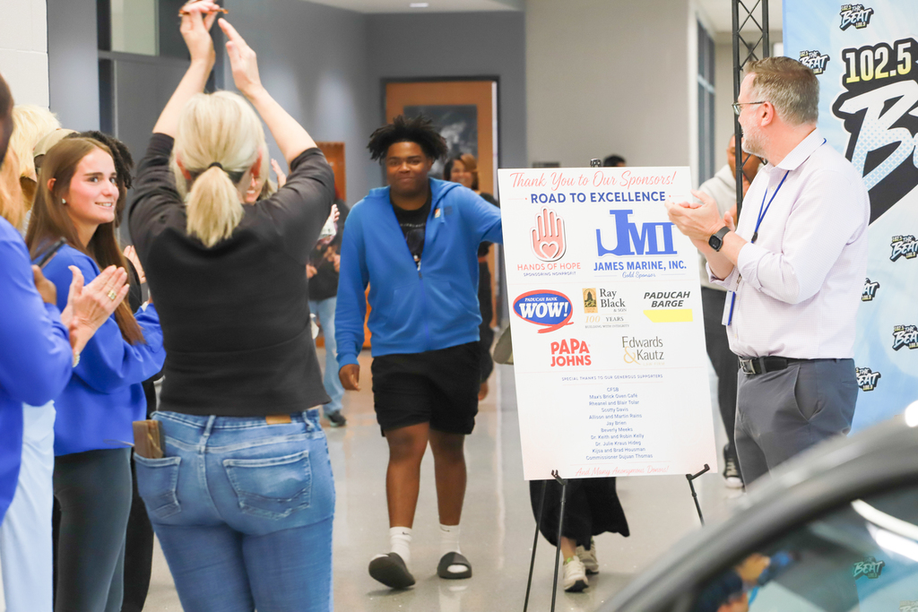 People standing around a sign in a hallway. One man claps, another walks away. Background includes banners and a car.