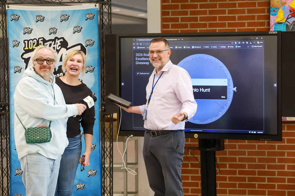 Two adults and a woman stand before a screen showing a video game. They hold a box.