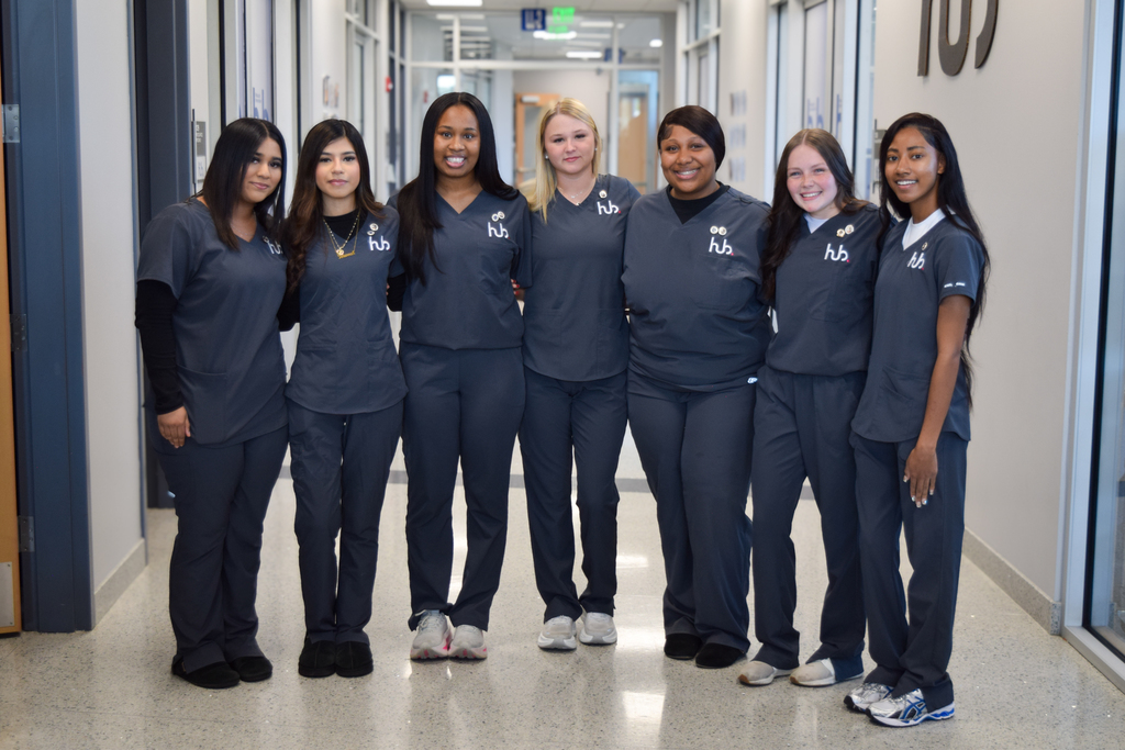 Seven women in matching uniforms stand in a hallway. The hallway has glass doors and gray flooring.